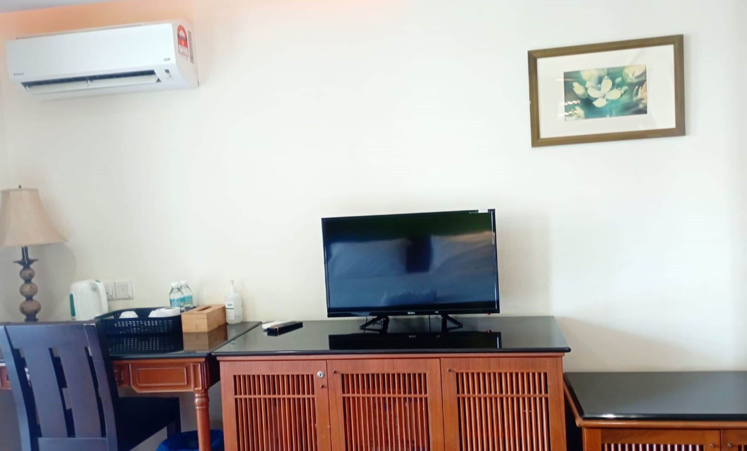 A room with a flat-screen TV on a wooden cabinet, a lamp, a bottle of hand sanitizer, and a framed picture on the wall.