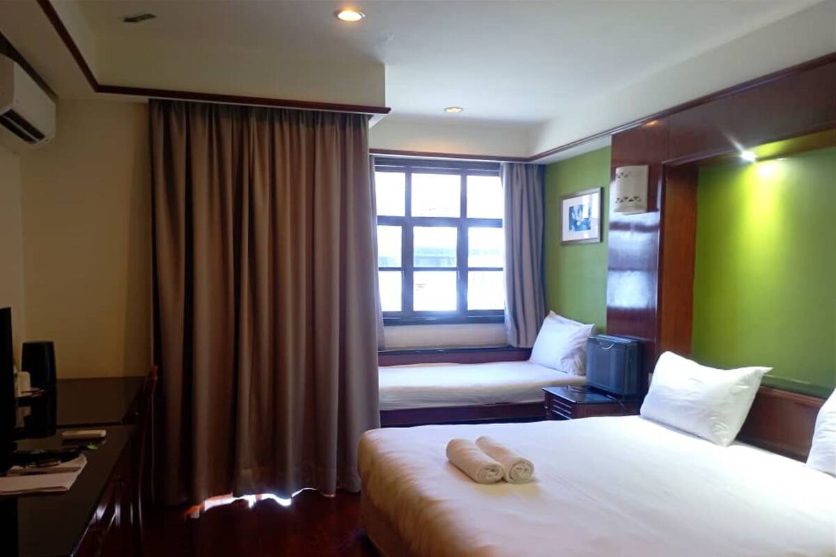 Quarto de hotel com uma cama grande, uma cama menor, uma escrivaninha com computador, janela com cortinas e parede verde.
