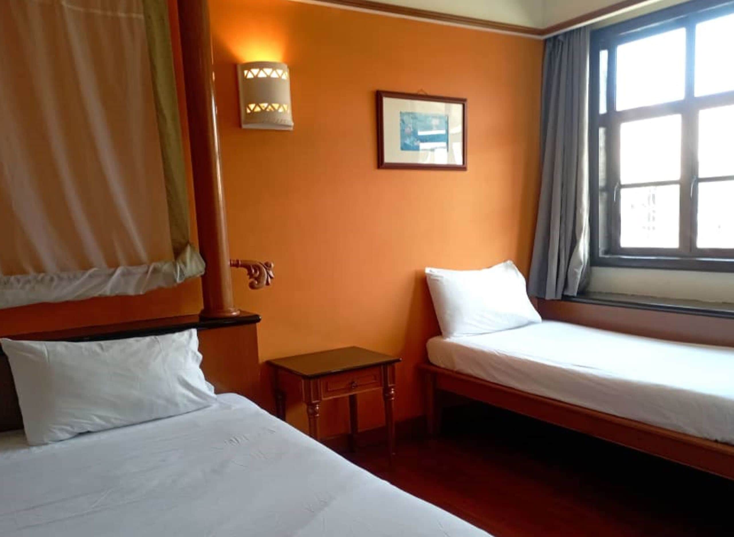 A hotel room with two single beds, a small table, a wall-mounted lamp, a framed picture, and a window with curtains.