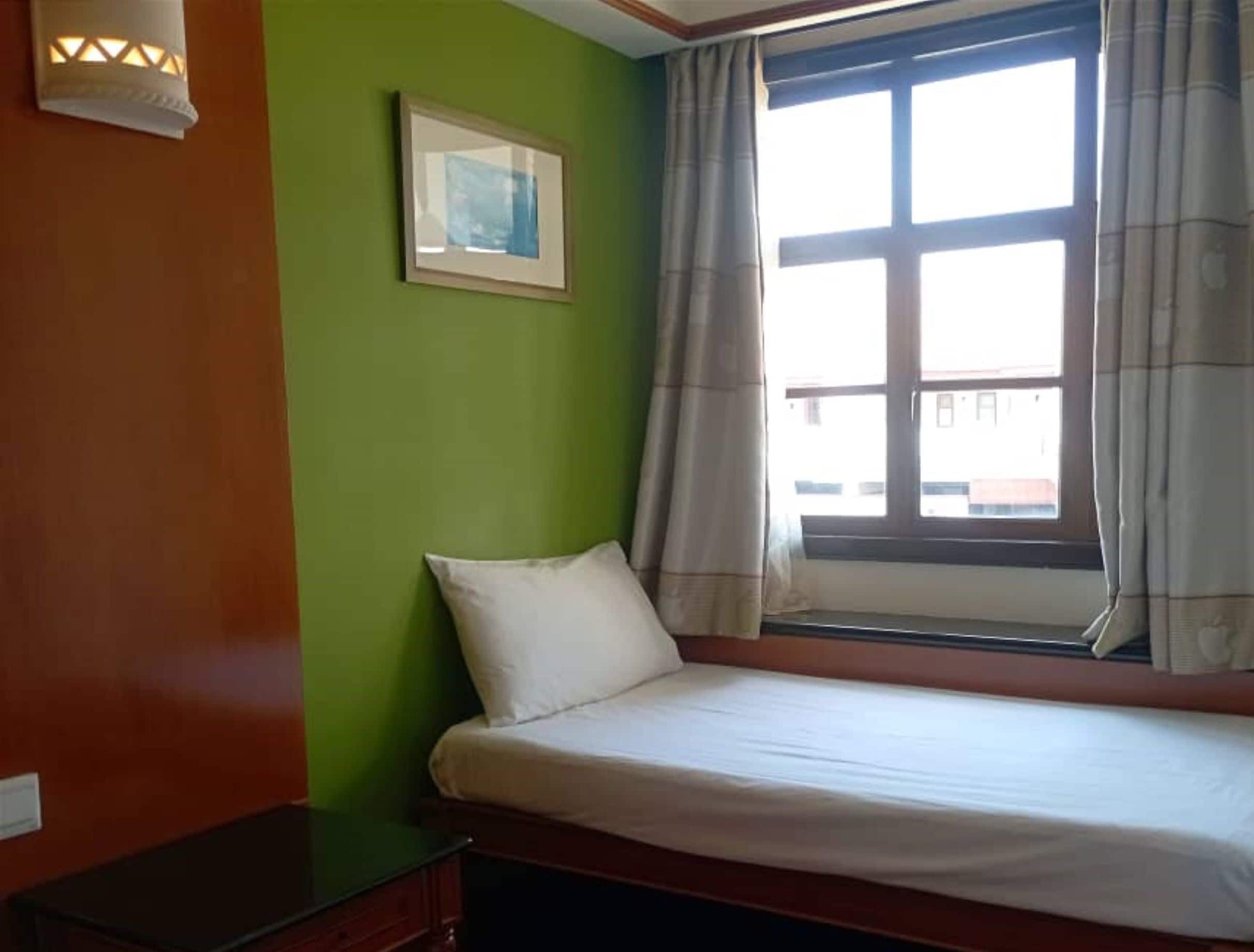 A small, single-bed room with a green wall, a framed picture, a window with curtains, and a bedside table.