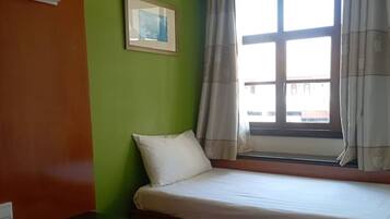 A small, single-bed room with a green wall, a framed picture, a window with curtains, and a bedside table.
