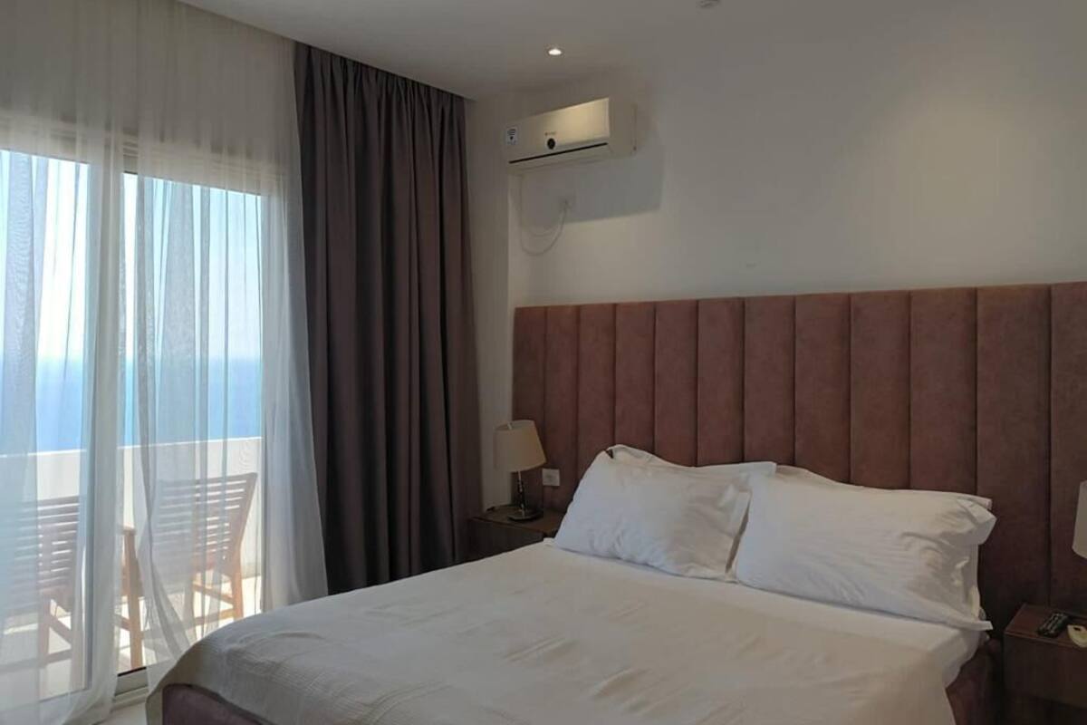 A hotel room with a large bed, a nightstand with a lamp, a wall-mounted air conditioner, and a view of the sea through sheer curtains.