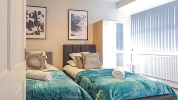 A hotel room with two beds, each with a teal bedspread and a white towel. There are two framed artworks on the wall above the beds.