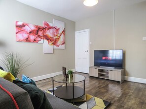 A living room with a flat-screen TV displaying 'NETFLIX', a glass coffee table, a sofa, and abstract wall art.