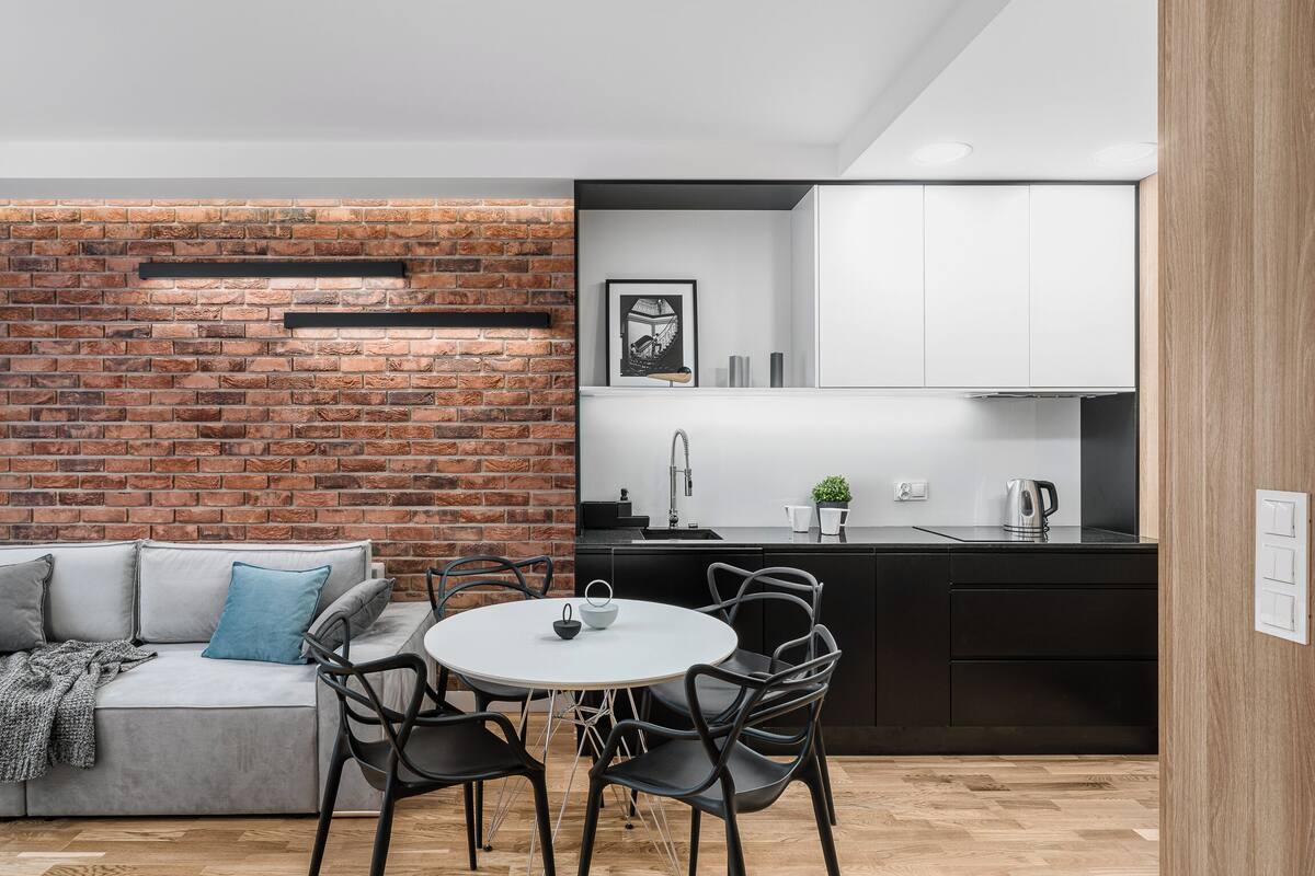 A modern kitchen with a brick wall, white cabinets, and a dining area with a round table and chairs.