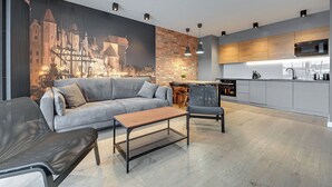 A modern living room with a grey sofa, a wooden coffee table, and a black chair. There is a kitchen area with grey cabinets and a microwave.