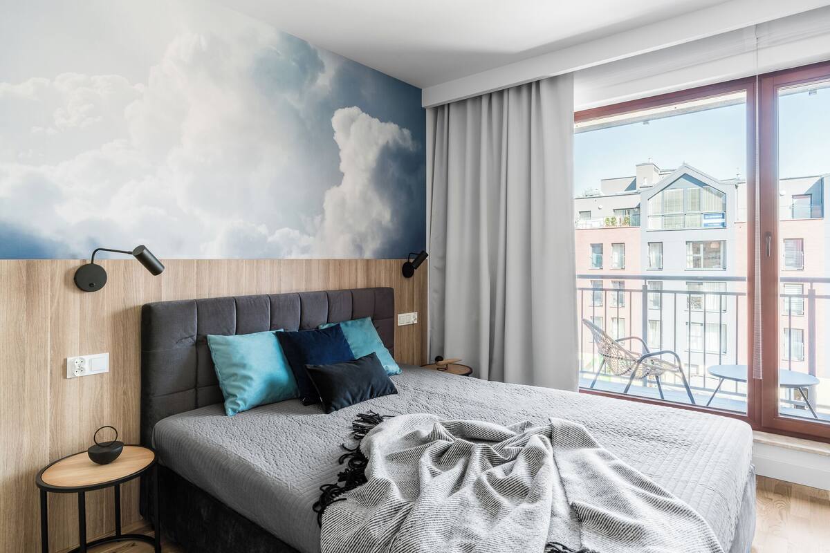 A modern hotel room with a large bed, a view of the city, and a cloud mural on the wall.