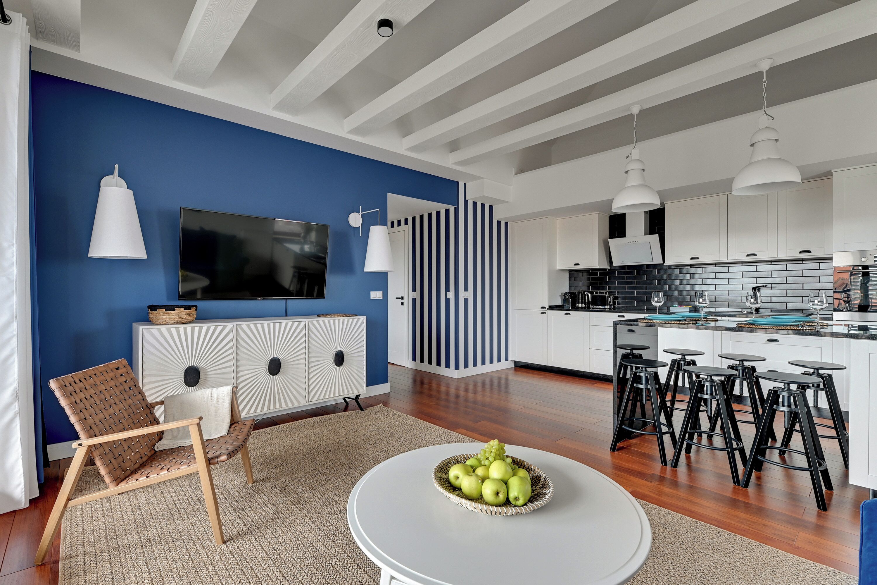 Un salon moderne avec un mur d’accent bleu, un téléviseur à écran plat, une table basse ronde sur laquelle est posé un panier de fruits, et un coin salon avec un fauteuil en osier.