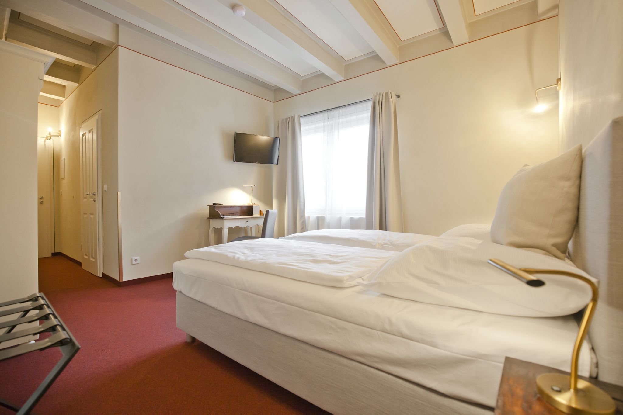 A hotel room with a bed, a TV mounted on the wall, a desk with a chair, and a window with curtains.