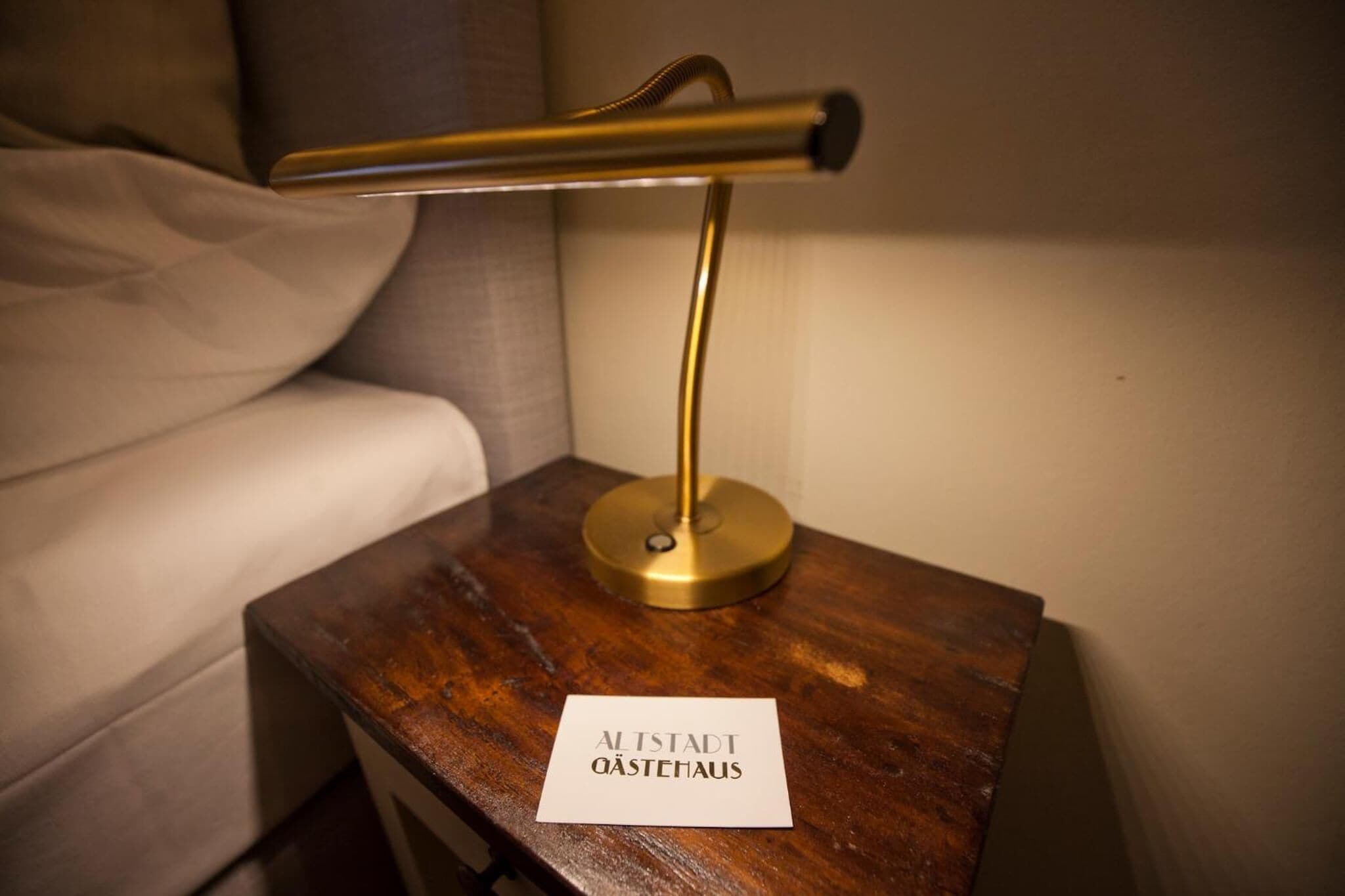 Uma mesinha de cabeceira com uma luminária de mesa em latão e uma placa com a inscrição 'Alstadt Qastehaus'.