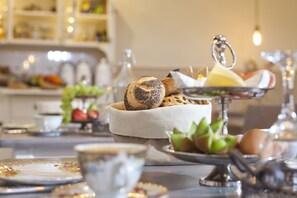 Een eettafel met verschillende soorten brood en gebak, een theepot en een schaaltje fruit.