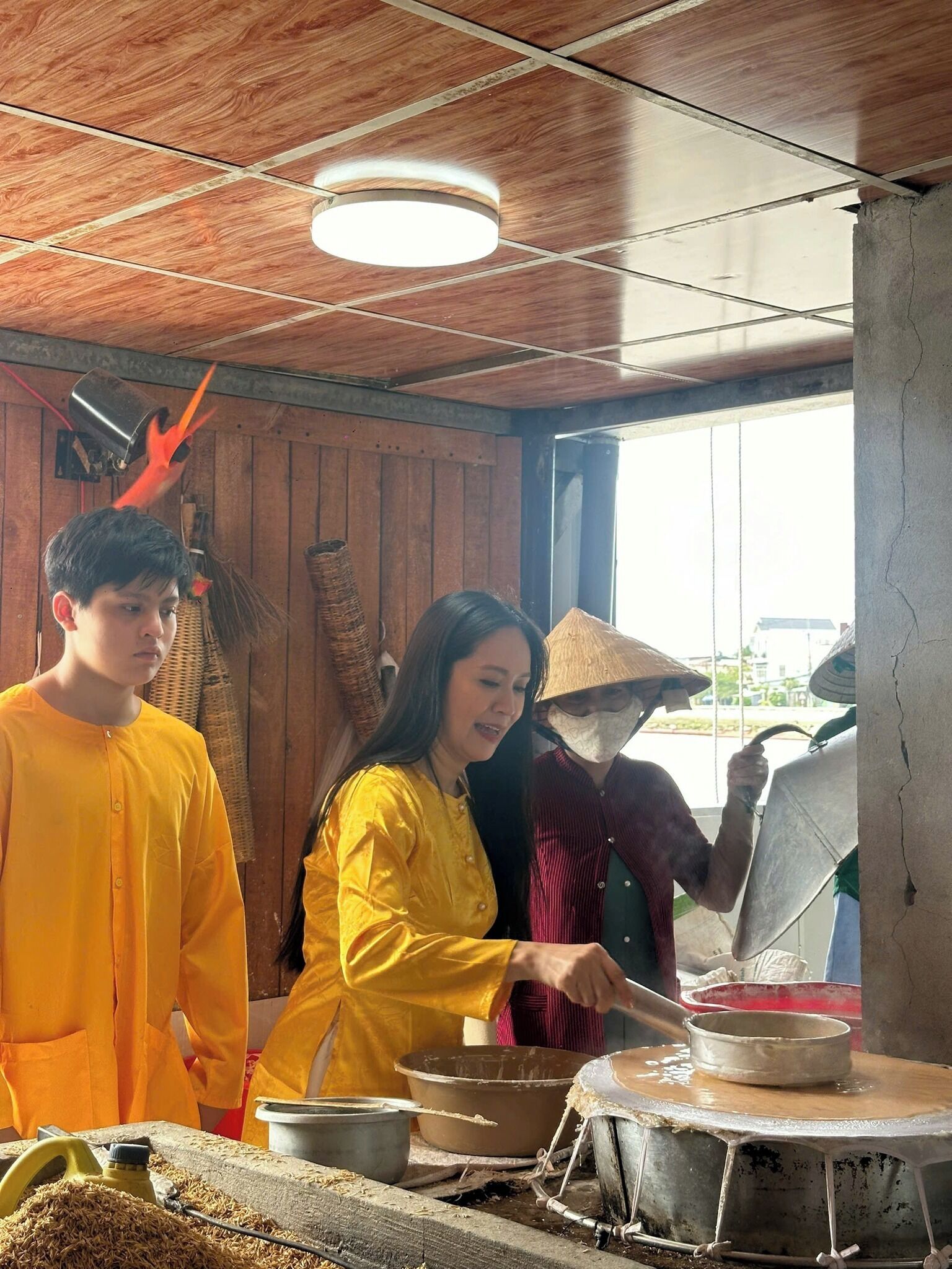 A traditional cooking setup with a woman preparing food, a man observing, and a person with a hat working nearby.