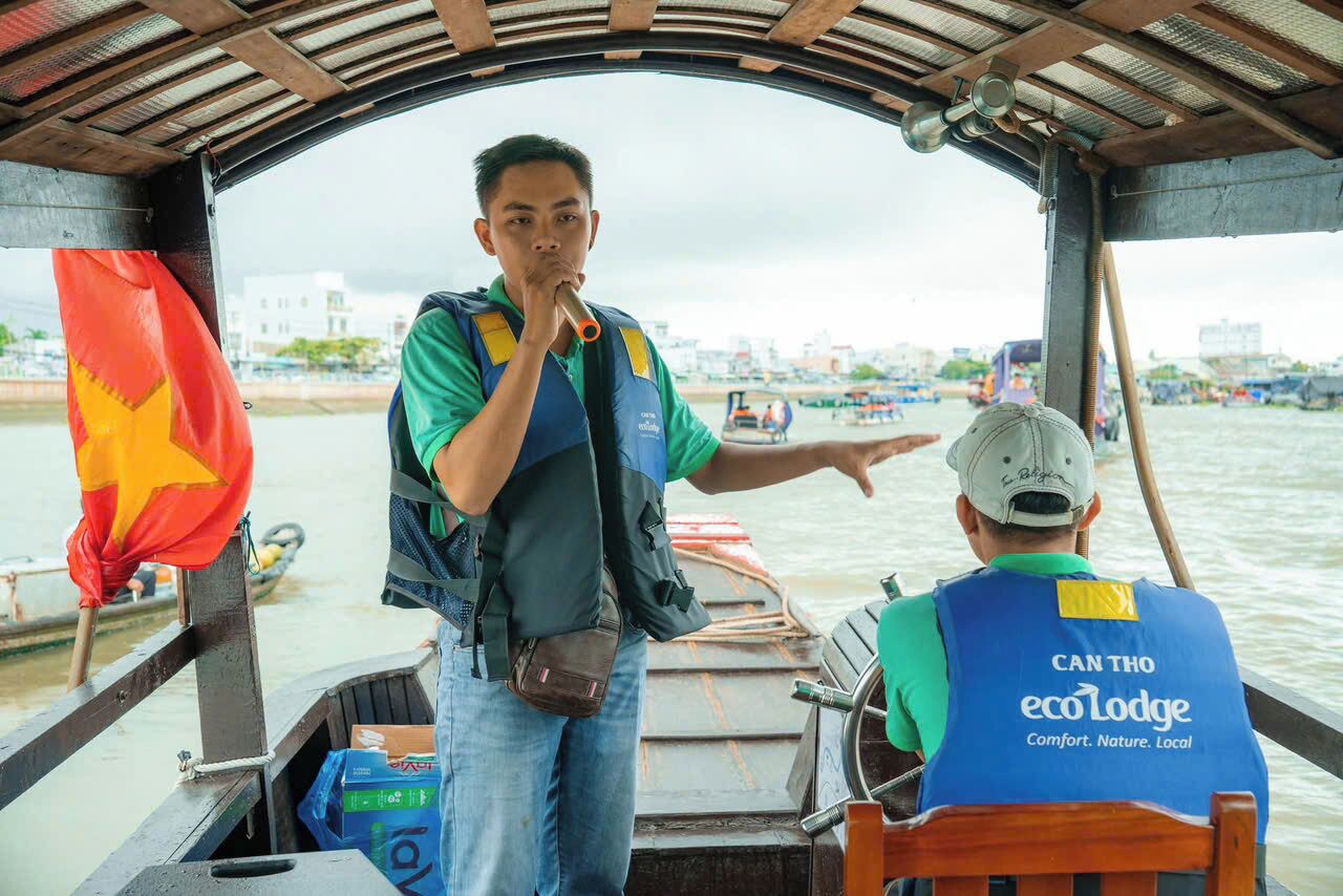 A boat interior with a person pointing towards the water, another person seated with a 'Cantho ecoLodge' vest, and a flag visible on the left.