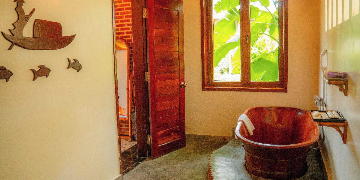 A bathroom with a wooden tub, a window with a view of green foliage, and a door leading to another room.