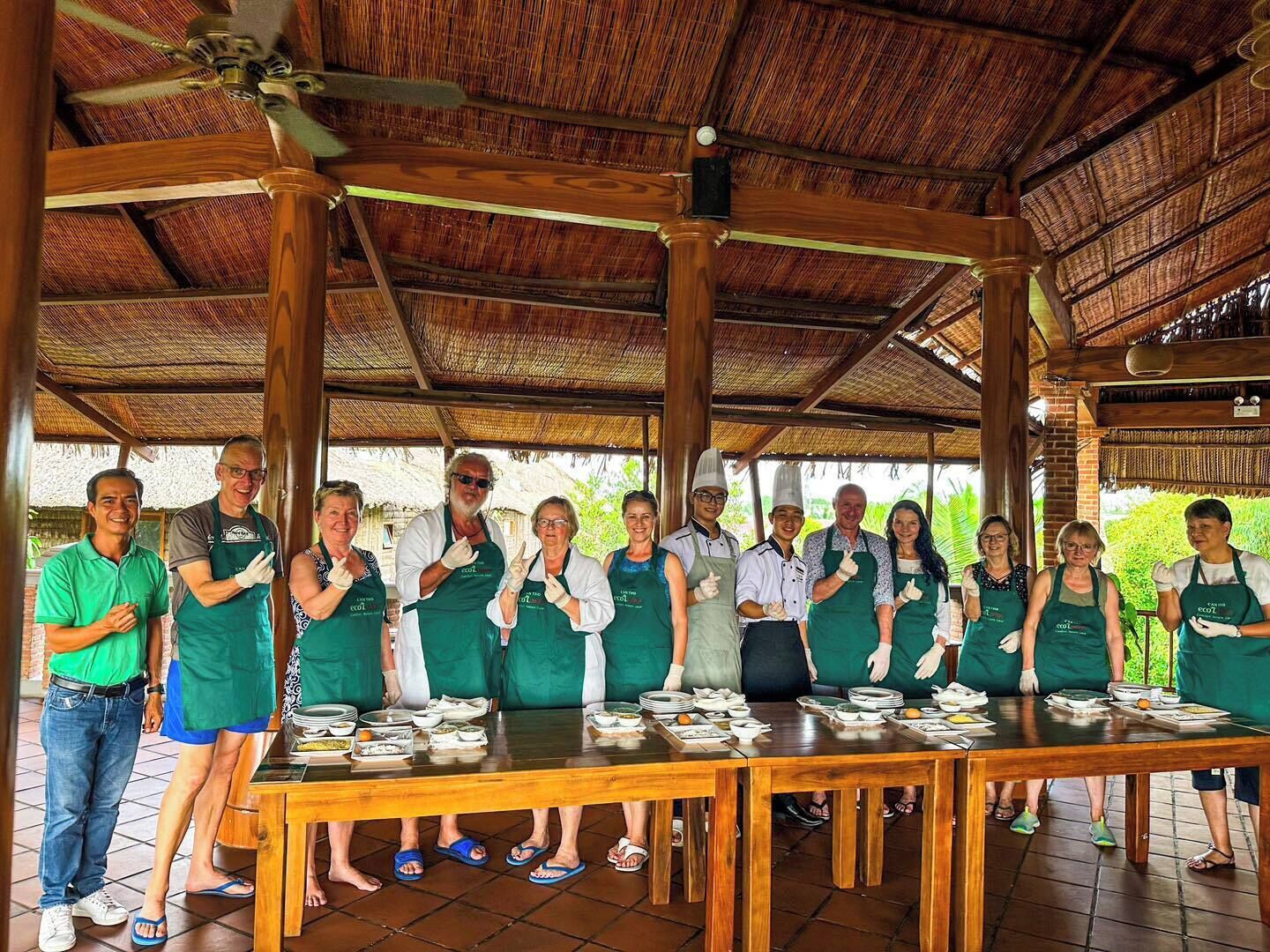 A group of people in aprons standing around a table with dishes, under a thatched roof structure.
