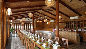 A long dining table set for a meal in a wooden-floored room with a bar area and a person at the bar. - Can Tho Ecolodge (Can Tho)