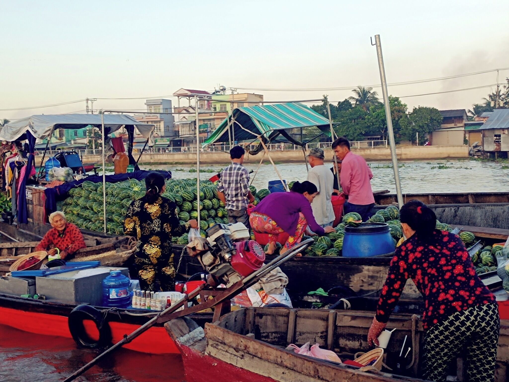 A group of people on a boat loaded with watermelons, with a cityscape in the background.