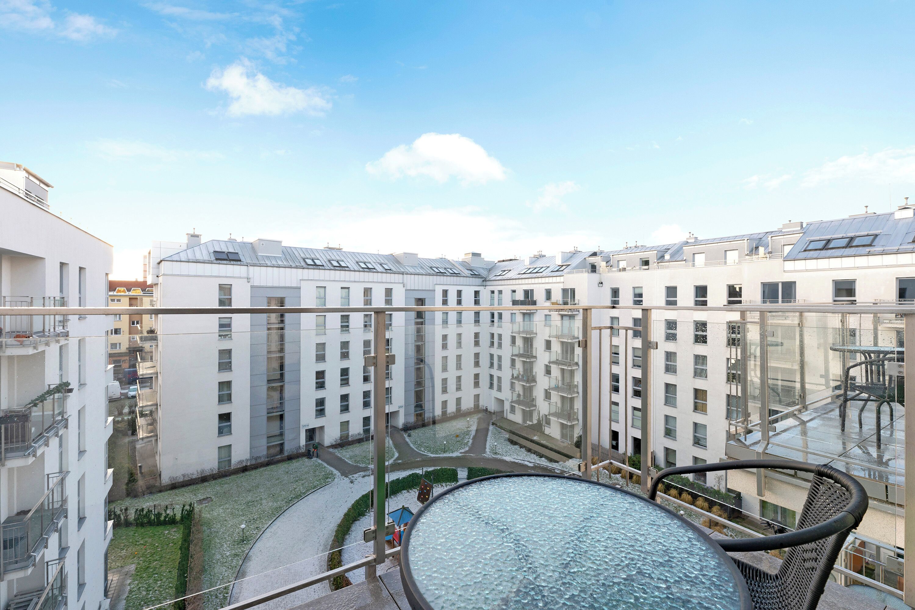 Un moderno complejo de apartamentos con varios balcones, barandilla de vidrio y un patio central con una mesa redonda y sillas.