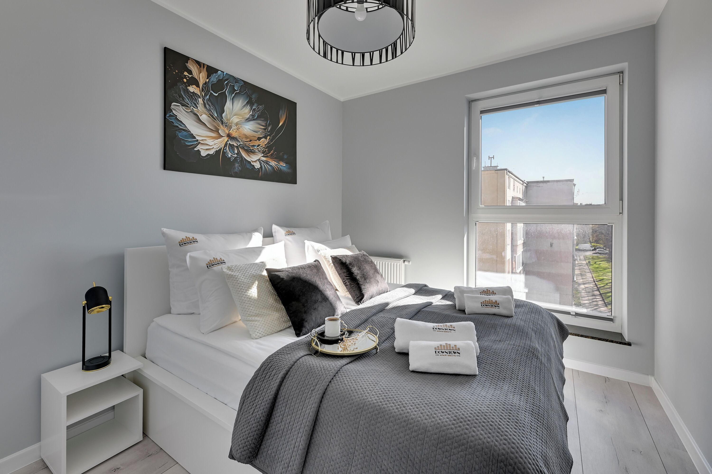 Een moderne slaapkamer met een groot raam, een bed met witte beddengoed, een decoratief kussen en een nachtkastje met een lamp.