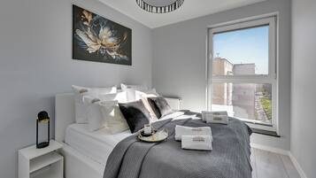 Een moderne slaapkamer met een groot raam, een bed met witte beddengoed, een decoratief kussen en een nachtkastje met een lamp.