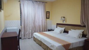 A hotel room with a bed, a desk, a chair, a nightstand, a telephone, and a window with curtains. - GREEN COURT HOTEL (Accra)