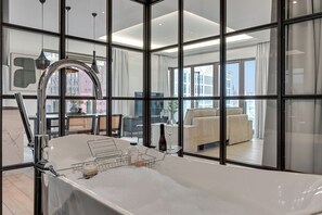 A modern bathroom with a bathtub, a glass shower enclosure, and a view of the living area through large windows. - Waterlane Riverside SPA, Pool & Gym by Downtown Apartments (Gdansk)