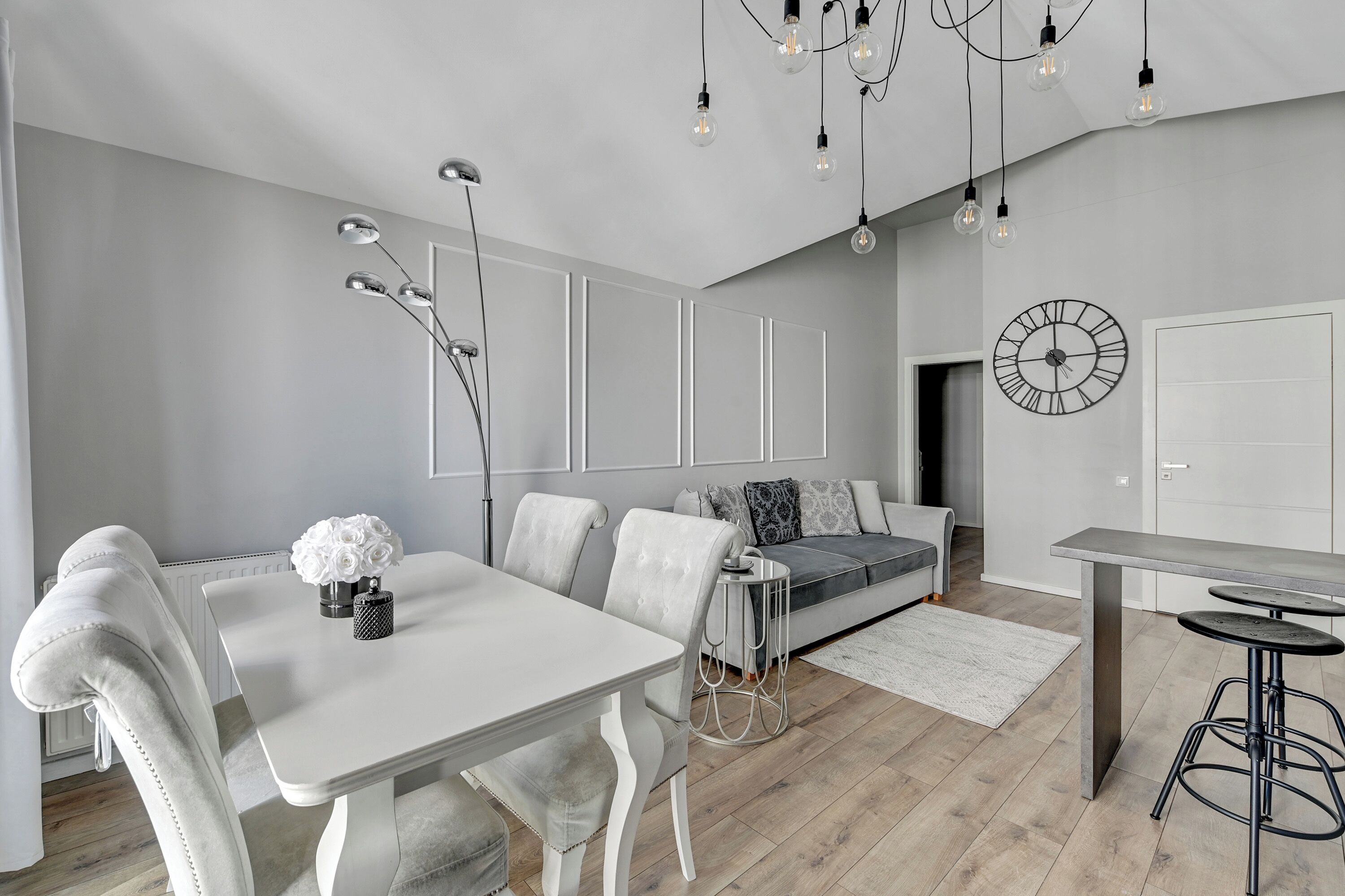 Ein modernes Wohnzimmer mit Esstisch, weißen Stühlen, einer Couch und einem Couchtisch. An der Wand hängt eine Uhr und über dem Tisch hängt ein Kronleuchter.