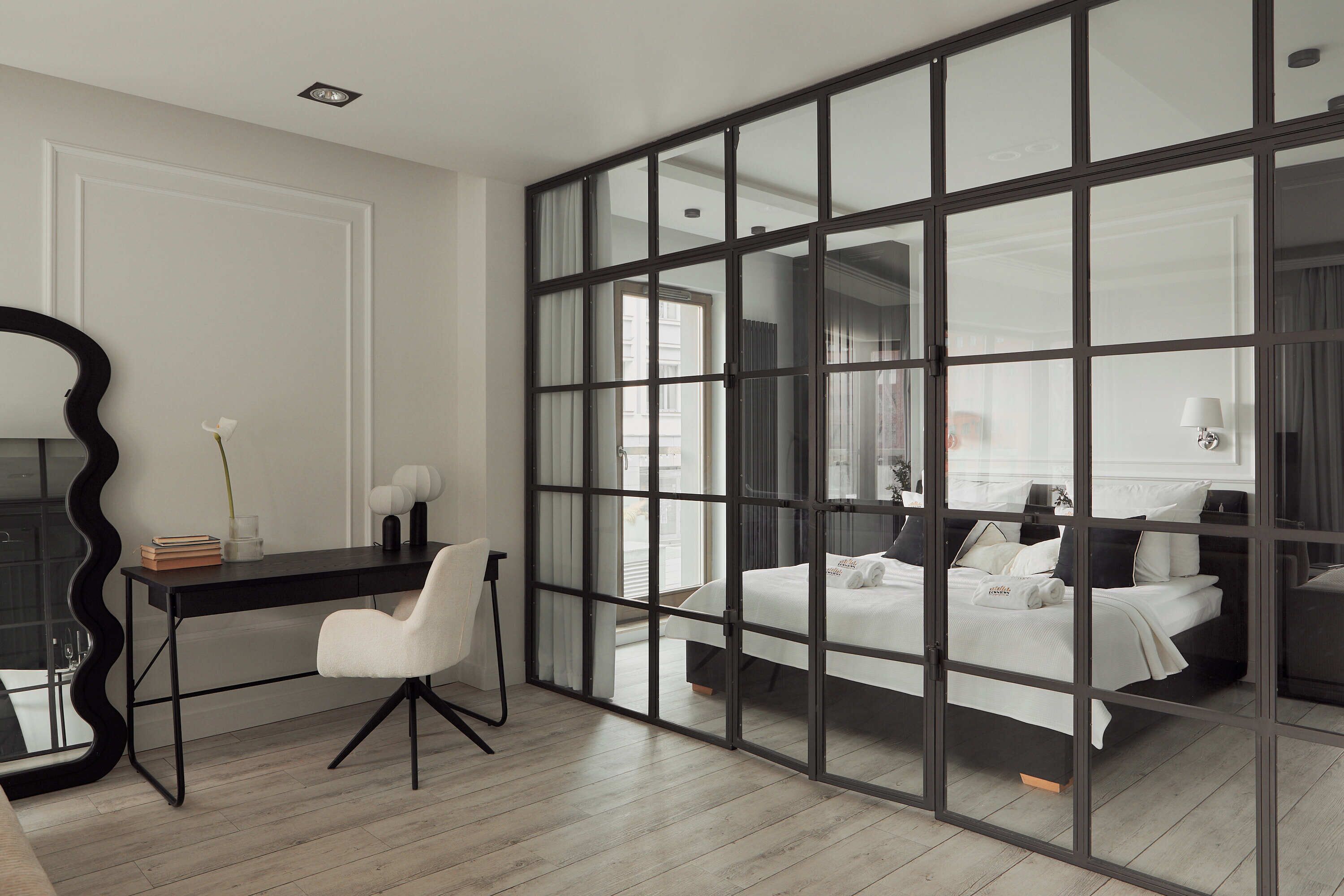 Habitación de hotel con un amplio ventanal corredero que da a un balcón, un escritorio con una silla, una cama con ropa blanca y un espejo.