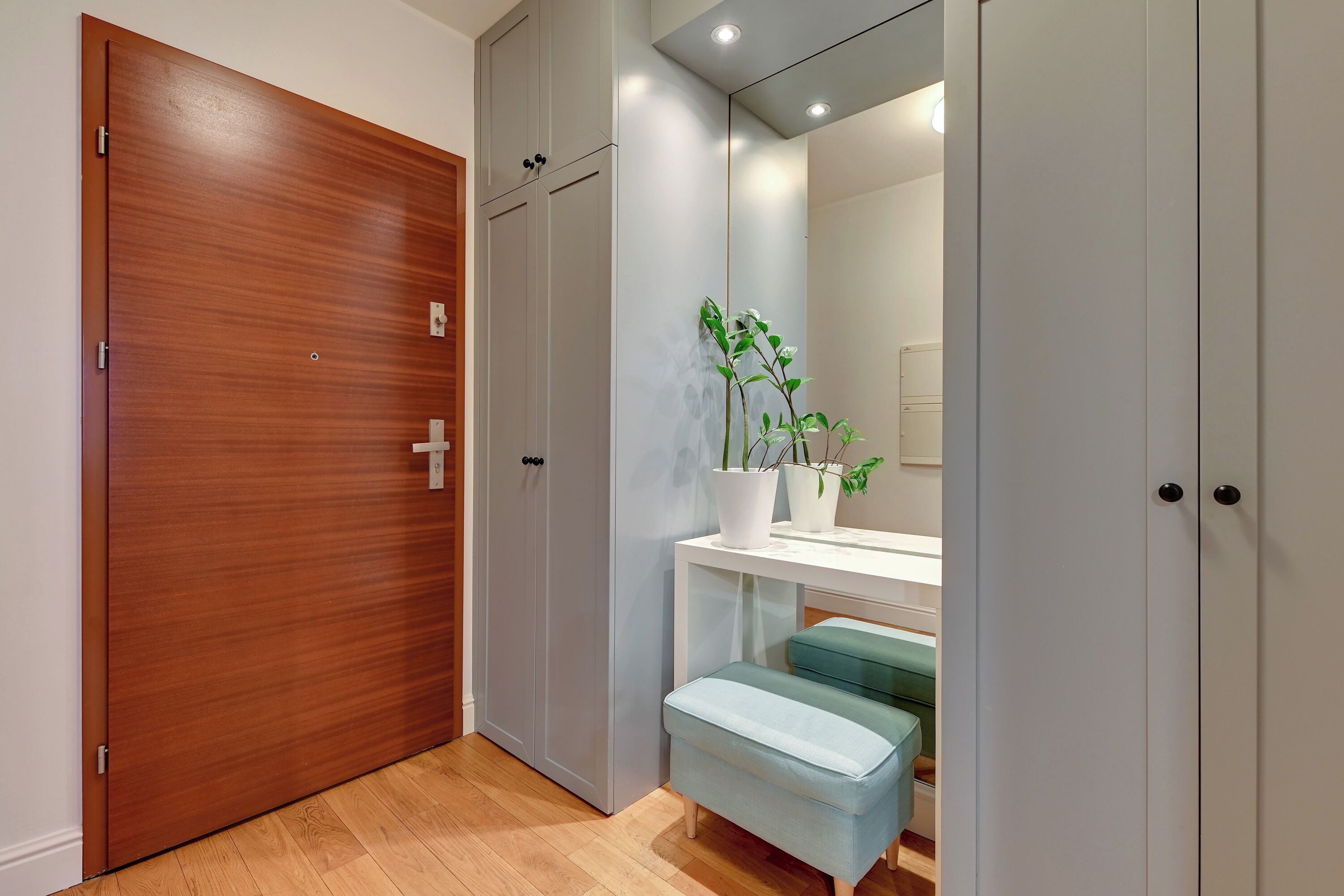 Un couloir avec une porte en bois, un banc blanc, une console blanche ornée d’une plante en pot et un miroir qui reflète une salle de bain.