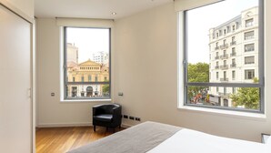 A modern hotel room with a large bed, a chair, and two windows offering city views.