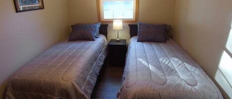 A small, narrow room with two single beds, a nightstand with a lamp, and a window with blinds.