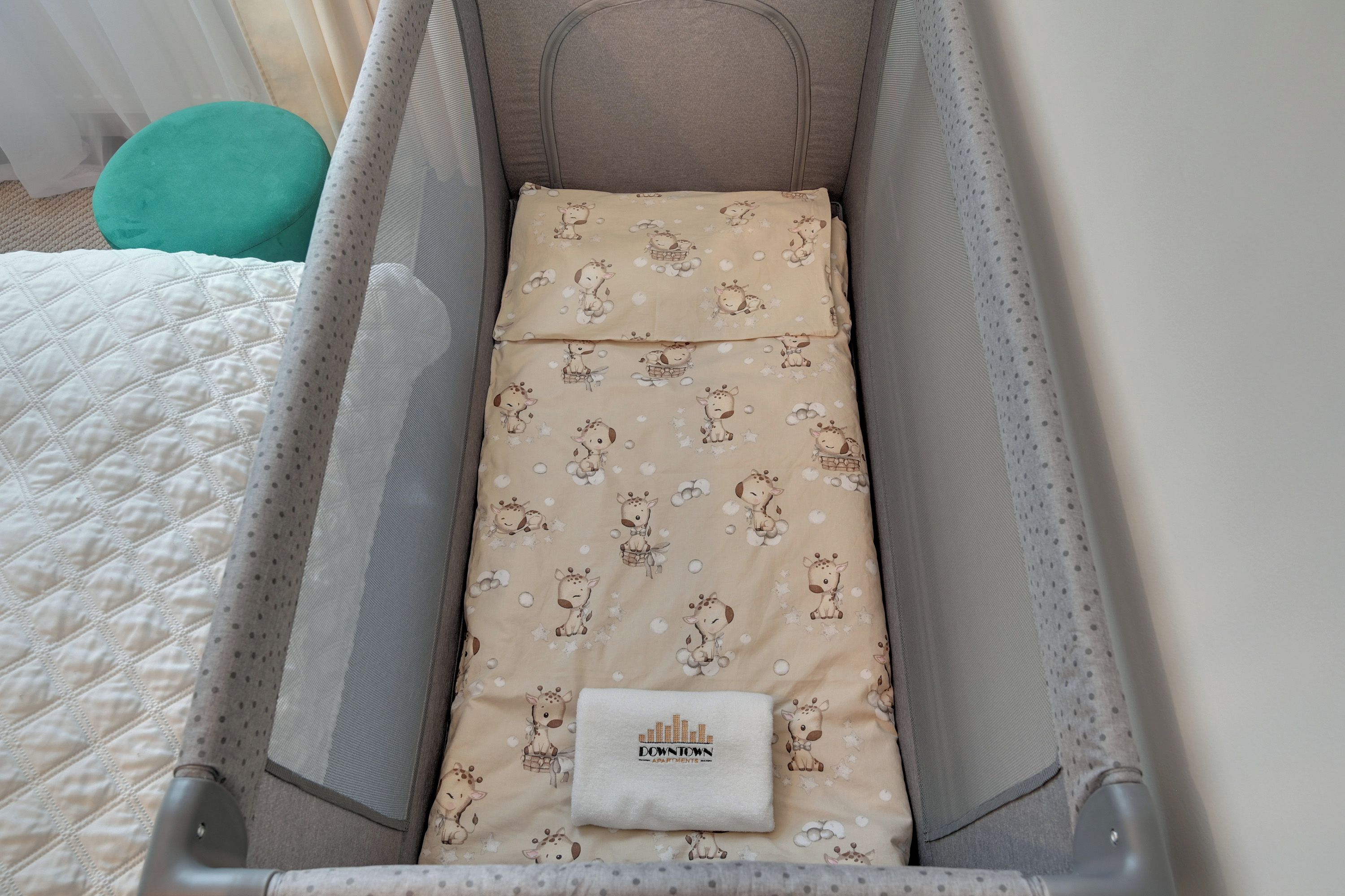A baby crib with a beige mattress featuring a giraffe pattern, a white pillow with 'Downtown' text, and a grey mesh side panel.