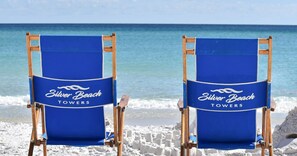 To blå strandstole med mærkaterne "Silver Beach Towers" på en sandstrand med havet i baggrunden.