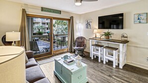 A living room with a sofa, a coffee table, a chair, a TV, and a balcony with a view of the building's exterior. - #121 BREAKERS, HILTON HEAD ISLAND, SC (Hilton Head Island)