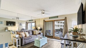 A spacious living room with a sofa, a coffee table, and a TV. There is a sliding glass door leading to a balcony with outdoor furniture. - #121 BREAKERS, HILTON HEAD ISLAND, SC (Hilton Head Island)
