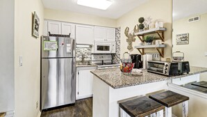 A compact kitchen with a refrigerator, microwave, stove, and toaster. There are two wooden stools and a small shelf with decorative items. - #121 BREAKERS, HILTON HEAD ISLAND, SC (Hilton Head Island)