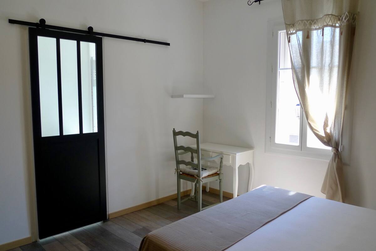 Ein Zimmer mit Bett, Stuhl, Schreibtisch, einer schwarzen Tür und einem fensterbepflanzten Fenster.