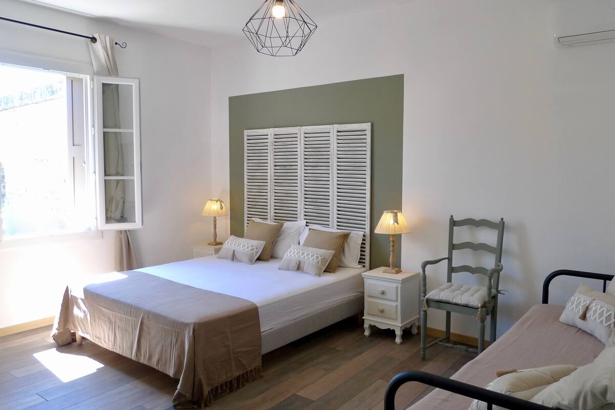 Ein Schlafzimmer mit einem Bett, zwei Nachttischen, einem Sessel, einem Fenster mit Vorhängen und einer geometrischen Leuchte.