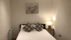 Ein ordentlich bezogenes Bett mit weißen Leinen und Karokissen, eine Nachttischlampe und ein an der Wand hängendes Bild.