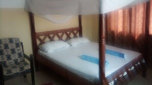 A single bed with a wooden frame and a canopy, a patterned chair, and a curtain in the background. - One World Apartments Bamburi (Mombasa)