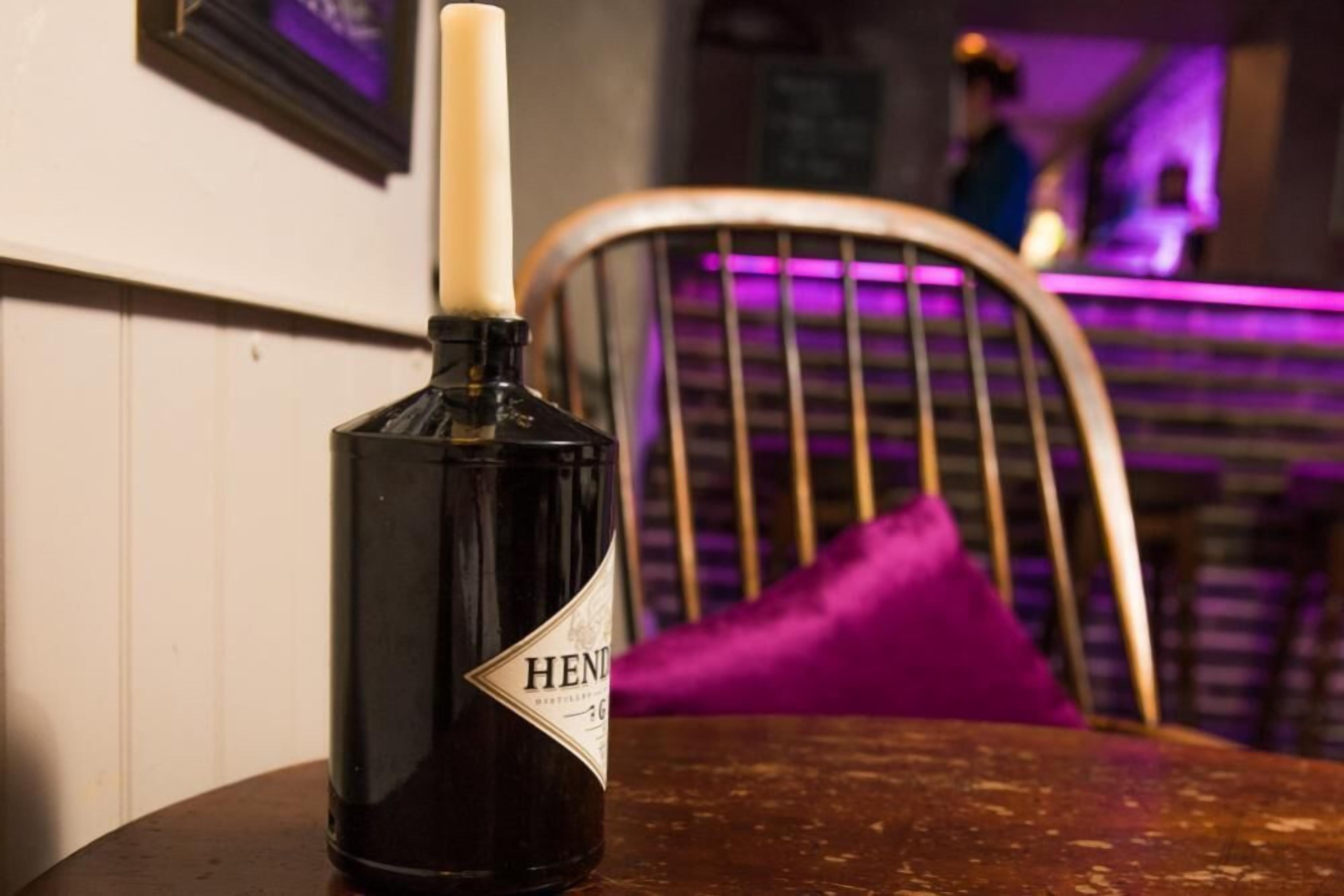 Una botella de cerveza Hendrick's sobre una mesa de madera con un cojín morado al fondo.