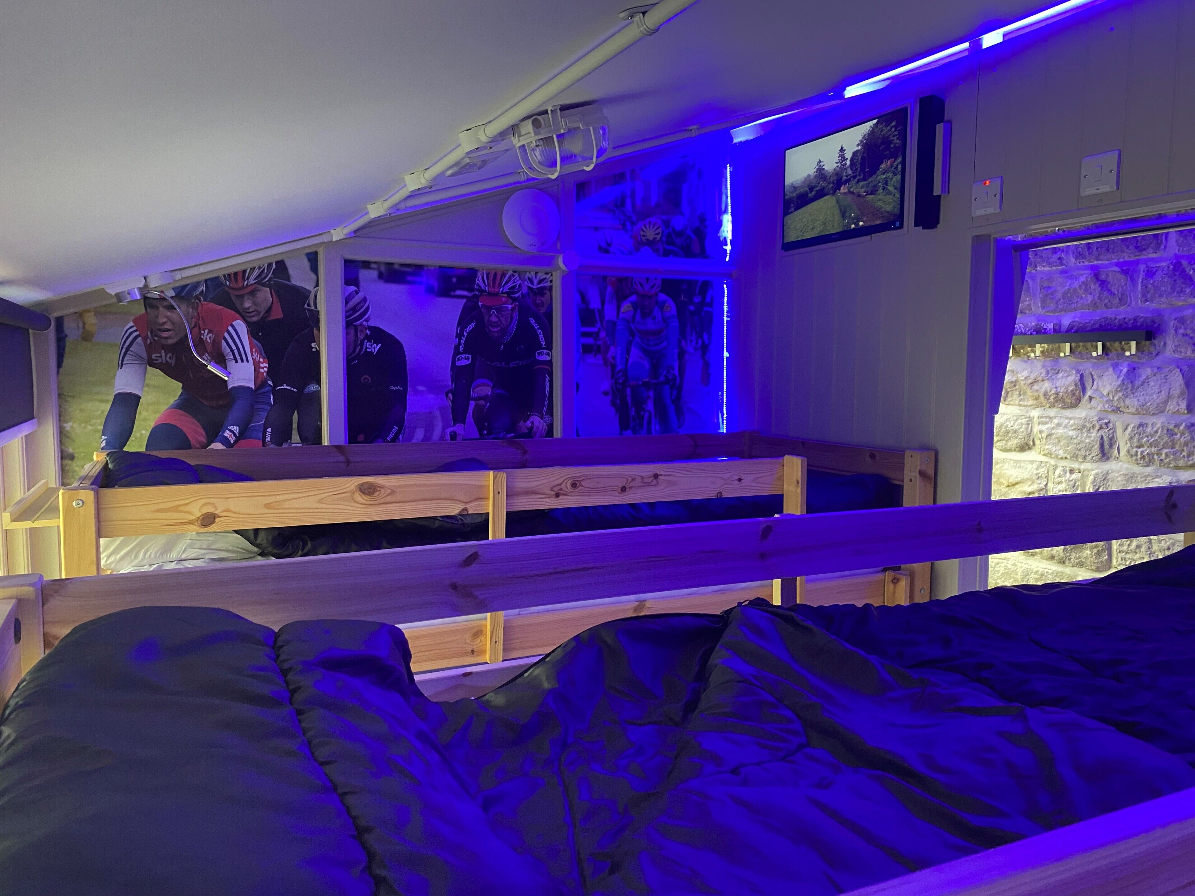 A bunk bed room with a television displaying a cycling event, a stone wall, and a ceiling-mounted projector.
