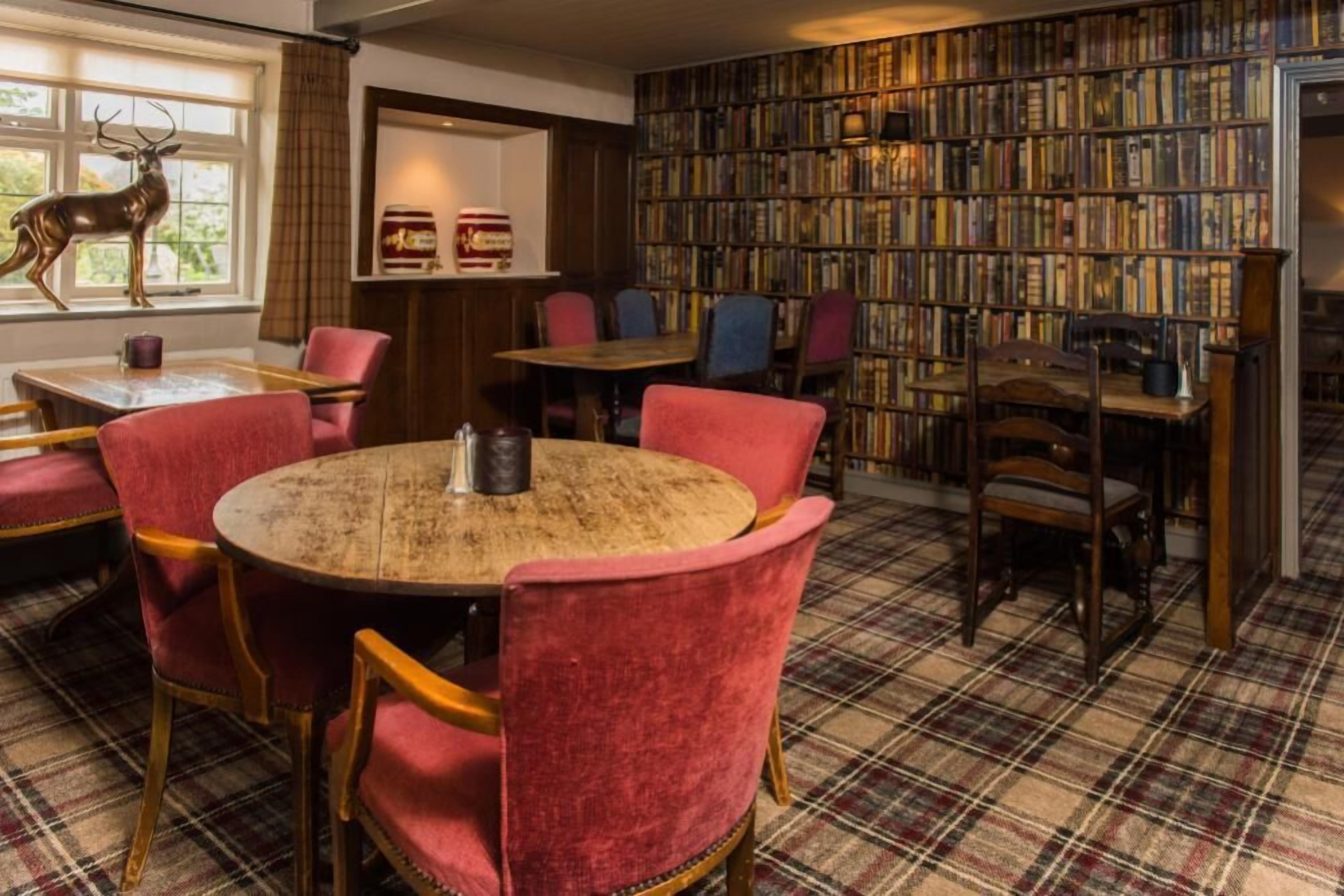 Een knusse bibliotheek met een ronde houten tafel, omringd door rode fauteuils. Een boekenplank vol boeken loopt langs de muur, en er staat een hertenbeeldje bij het raam.