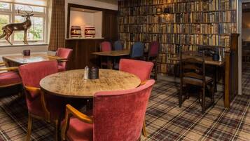 Een knusse bibliotheek met een ronde houten tafel, omringd door rode fauteuils. Een boekenplank vol boeken loopt langs de muur, en er staat een hertenbeeldje bij het raam.