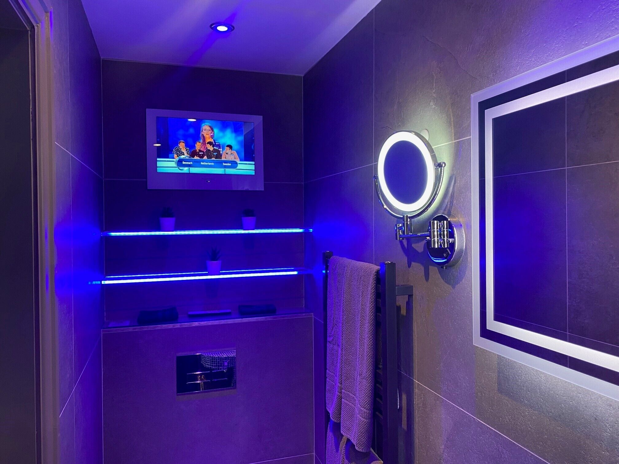A modern bathroom with a TV mounted on the wall, a vanity with a mirror, and blue LED lighting.