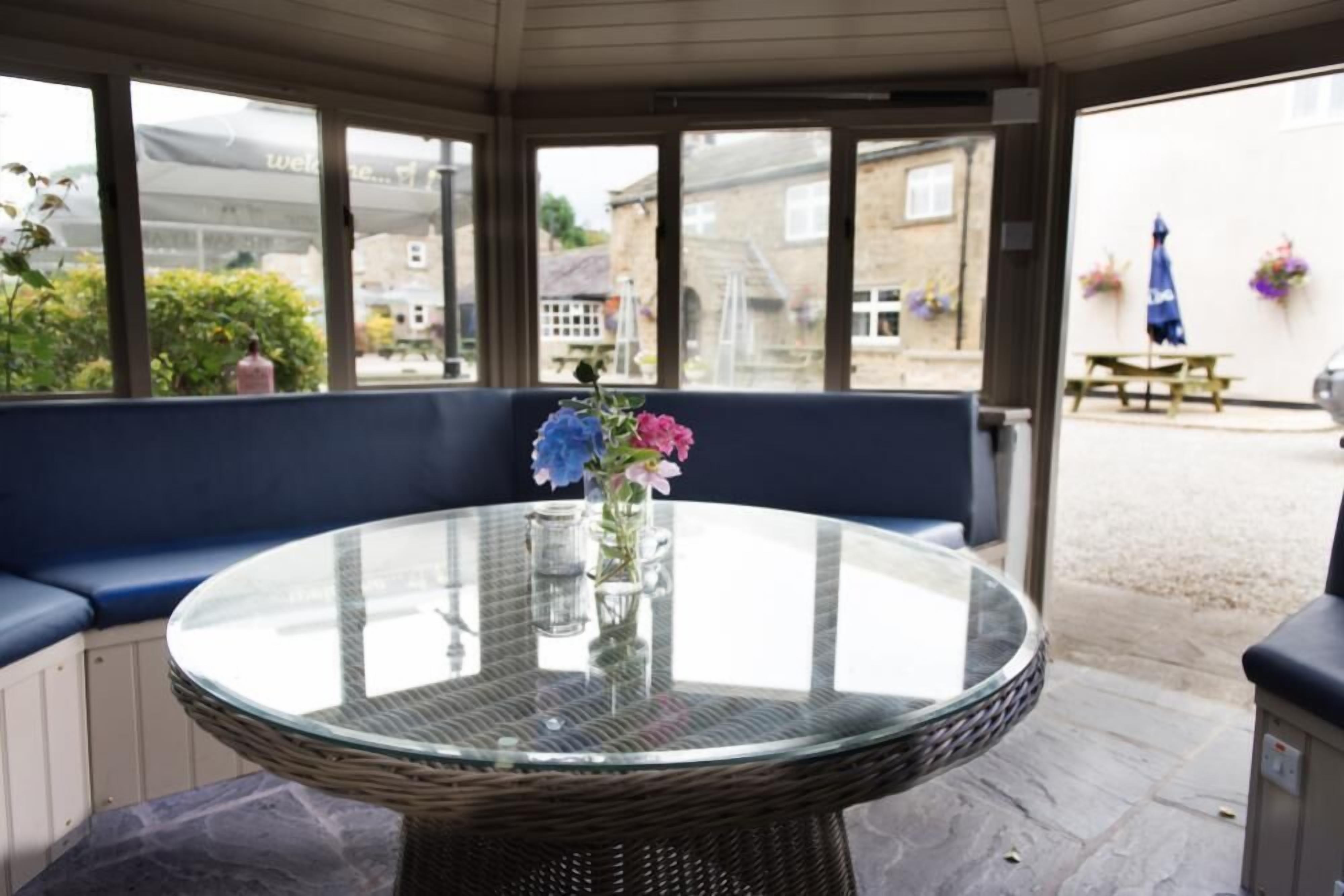 A glass-top table with a wicker base, a blue cushioned bench, and a vase with flowers.