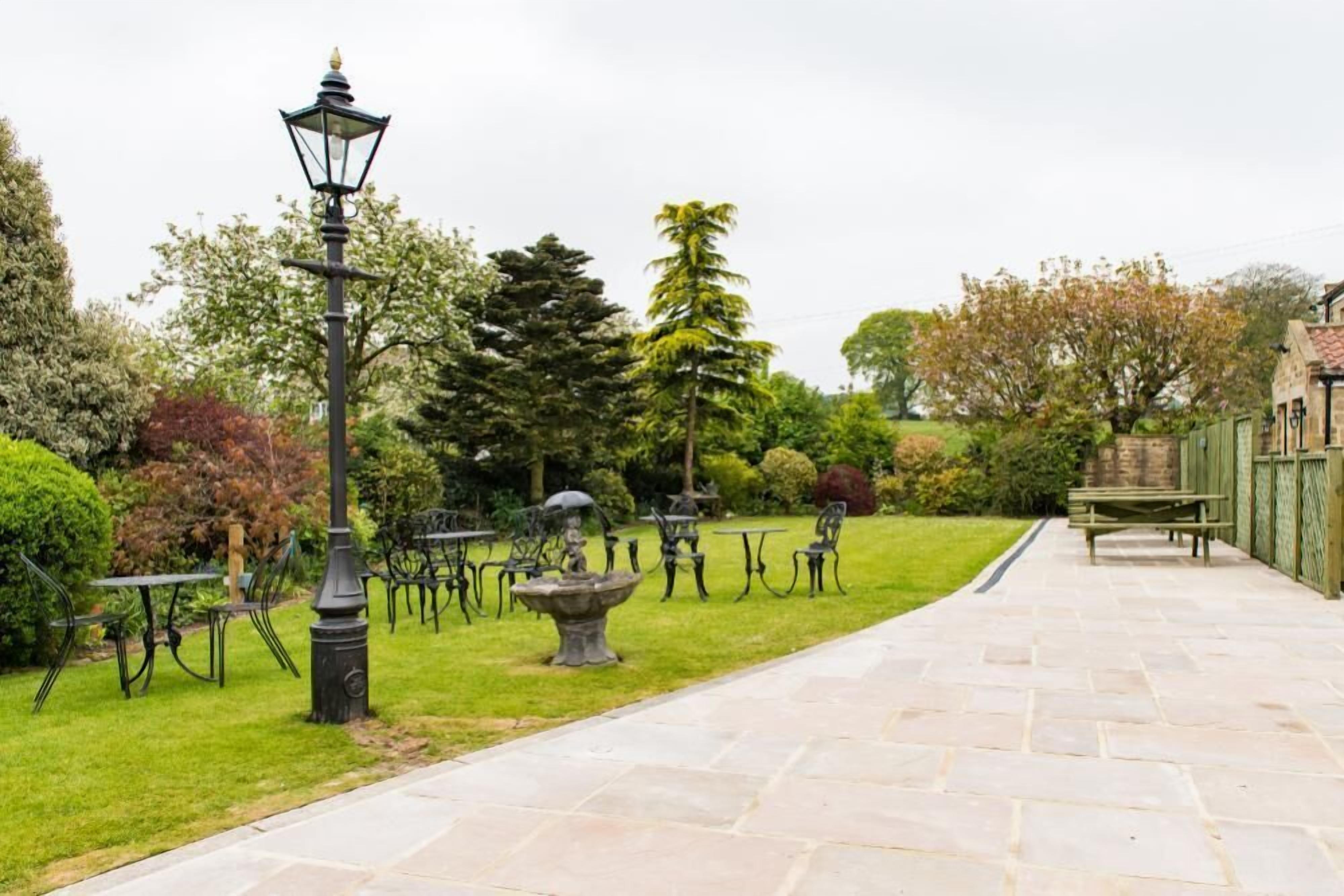 Un jardin avec des allées pavées de pierres, des tables et des chaises en métal, et un lampadaire.