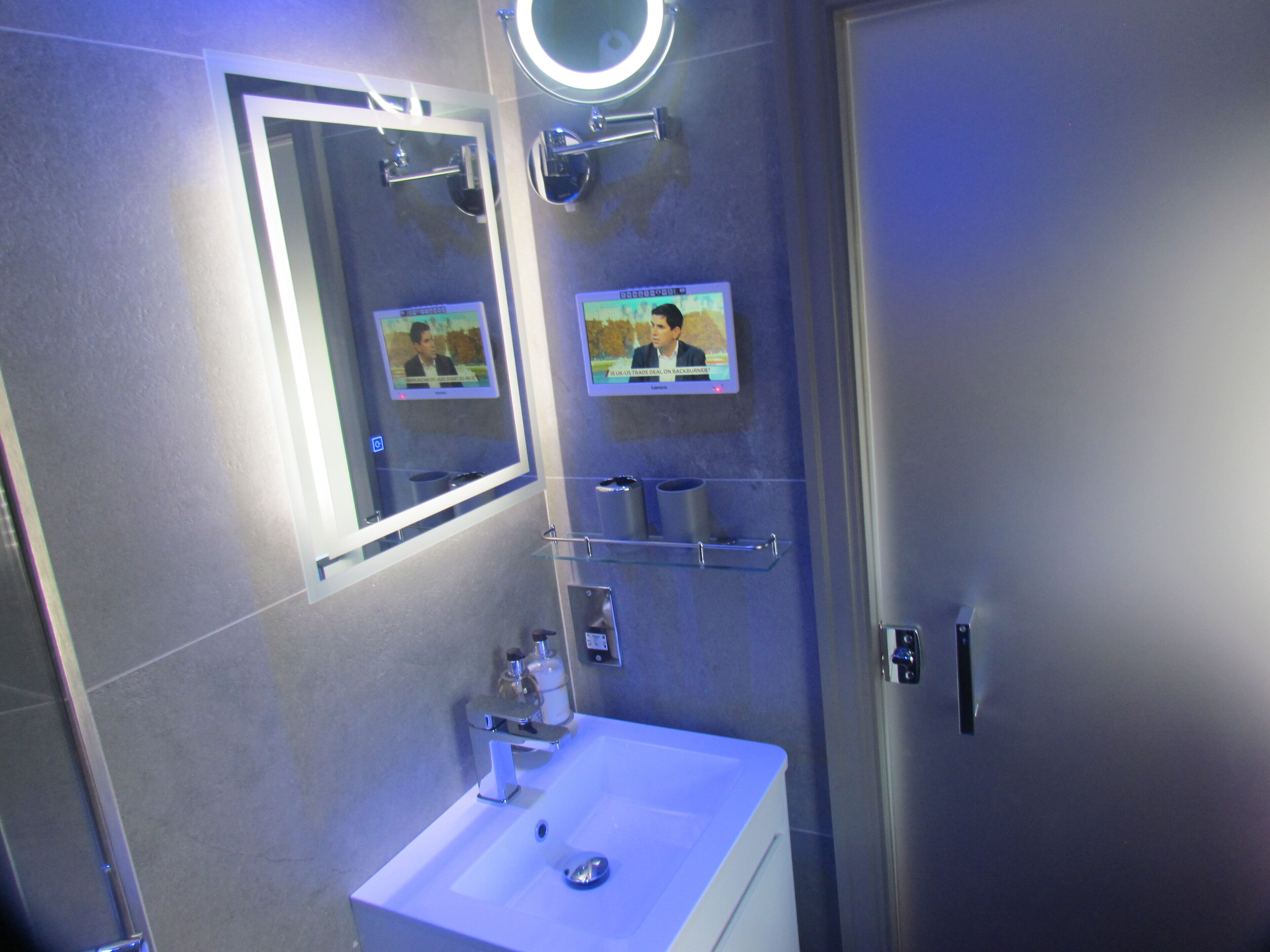 Un baño moderno con un lavabo blanco, un espejo con luz integrada y dos pantallas mostrando noticias.