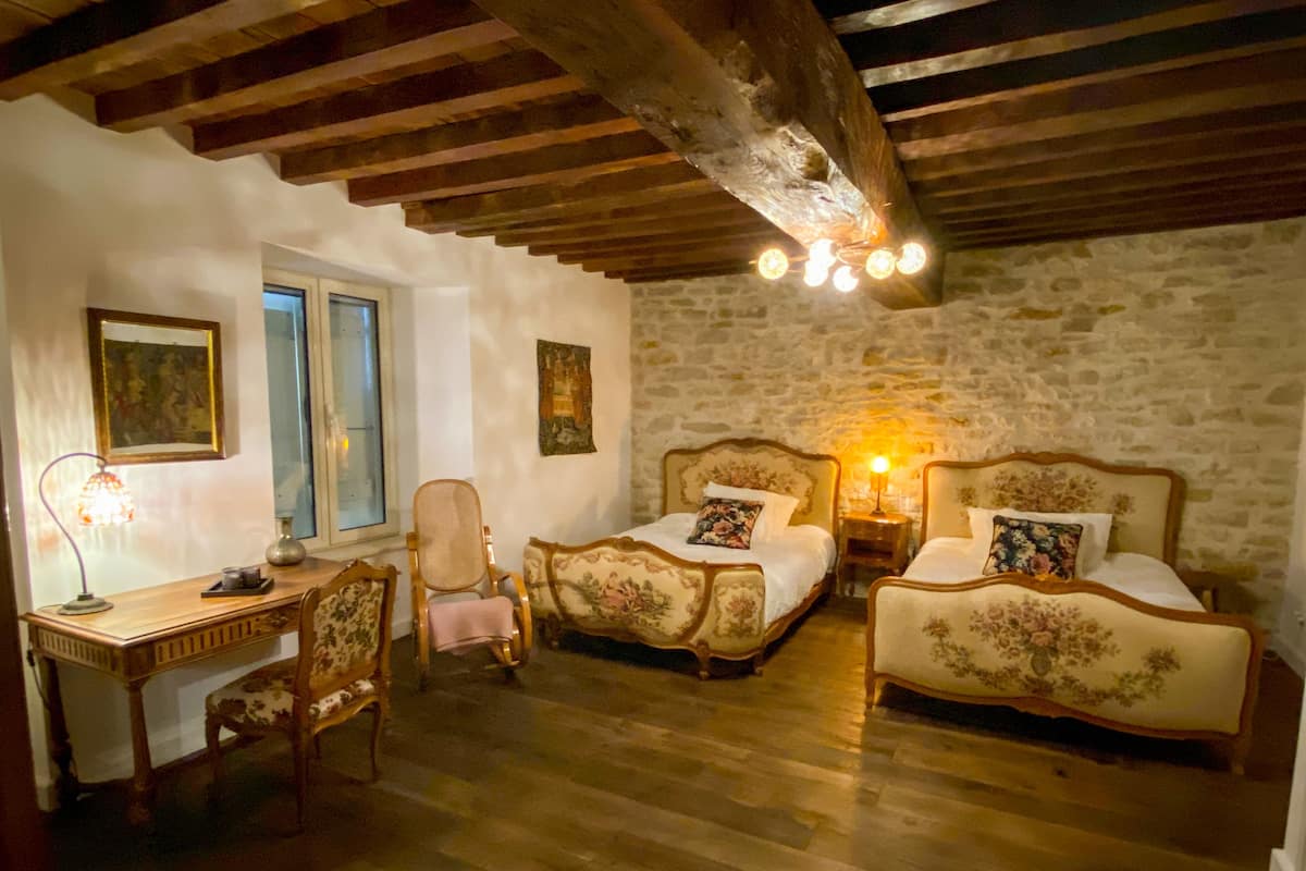 A bedroom with two beds, a desk, and a chair. The room has wooden beams on the ceiling and stone walls.
