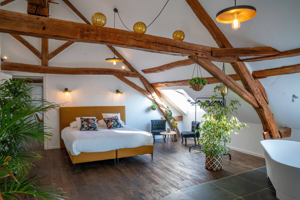 A bedroom with a wooden ceiling, a bed, a chair, a small table, and a bathtub.
